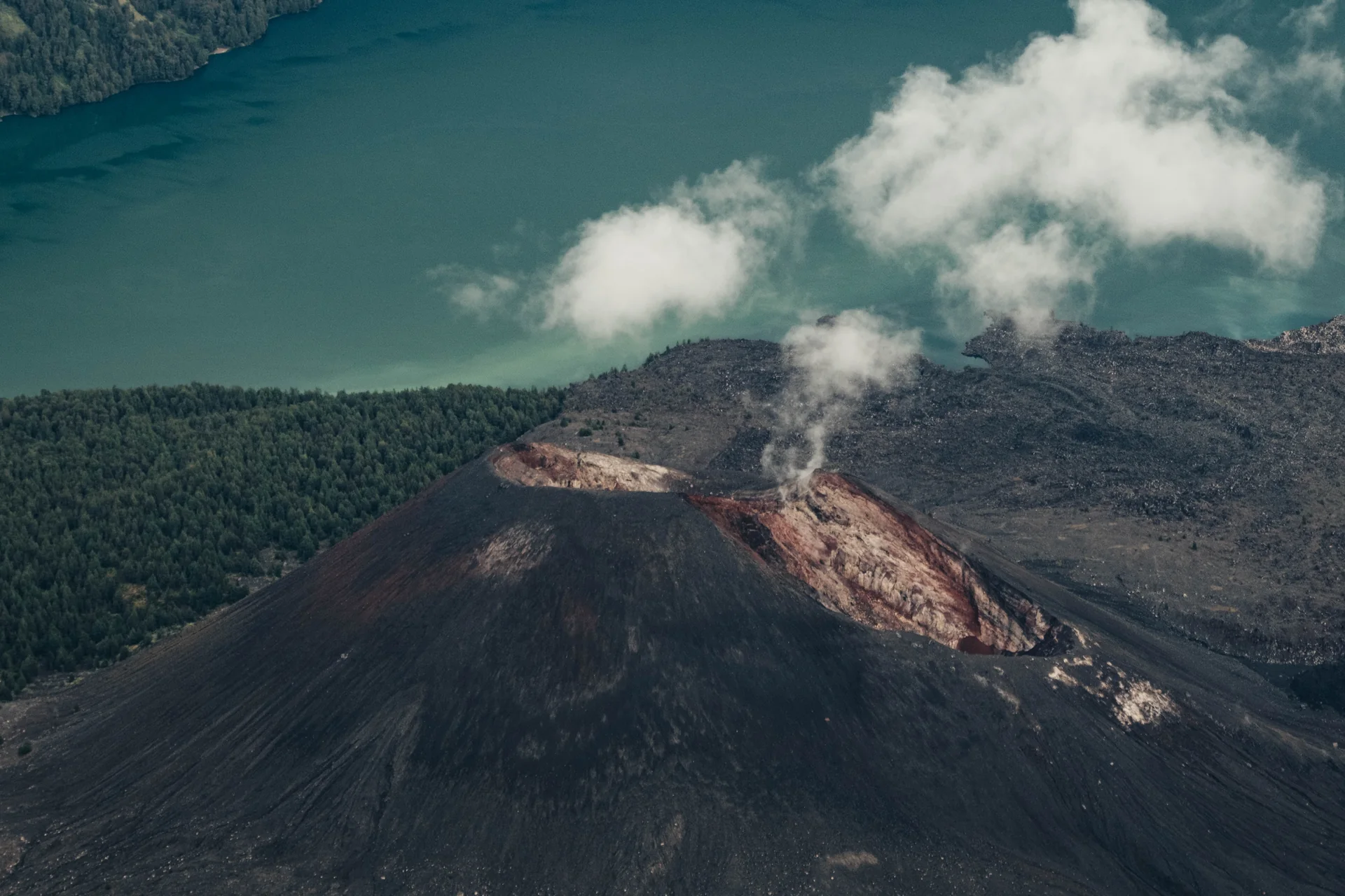Volcanic cone with a plume beside a turquoise lake